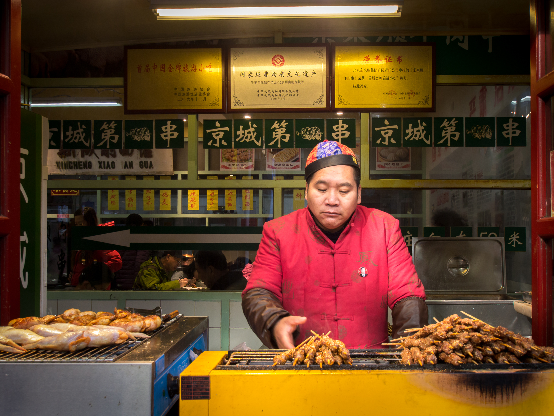 A Beijing street-food seller in a red jacket standing behind trays of skewers